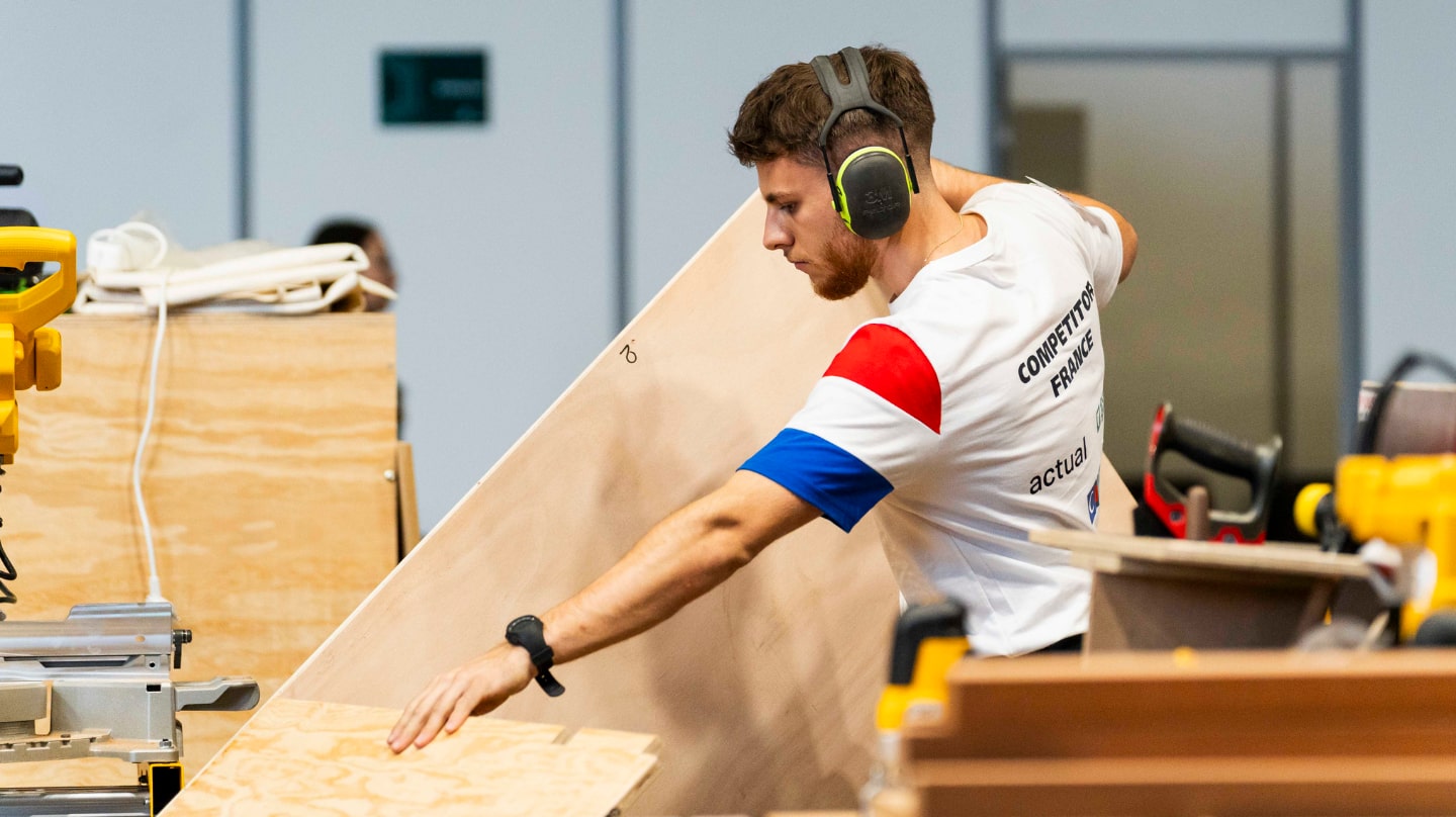 Un jeune homme avec un tee-shirt avec un drapeau tricolore manipule du bois. Il porte un casque anti-bruit.